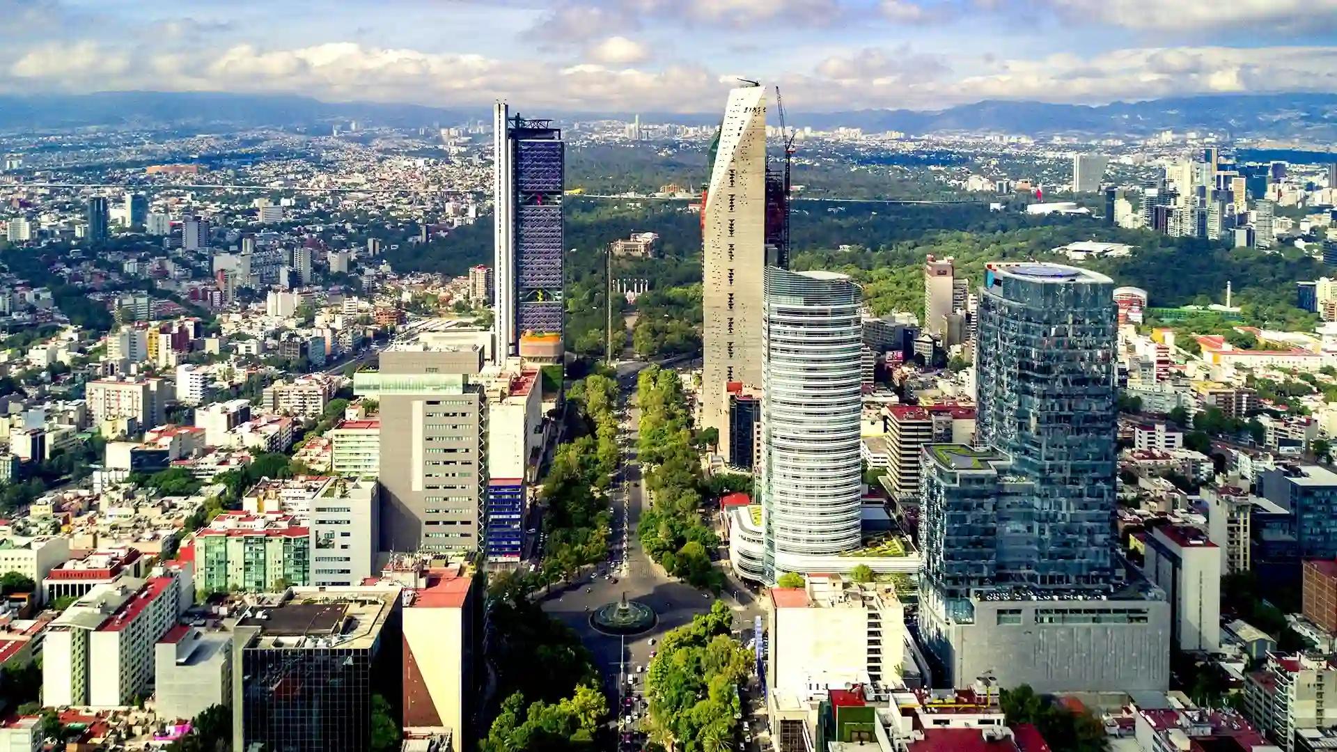 Modern skyline of Mexico City with tall glass skyscrapers, tree-lined streets, and a landmark roundabout at the center.